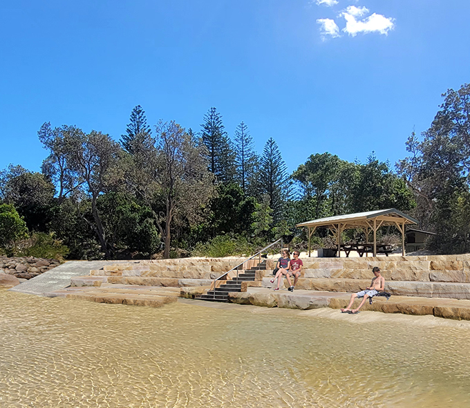 Beach scene with individuals seated on stone steps leading to the water, under a clear blue sky