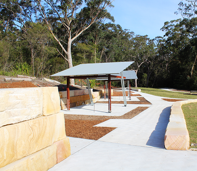 A shelter with a metal roof supported by steel pillars, adjacent to a pathway lined with large stone blocks and wood chips, surrounded by greenery