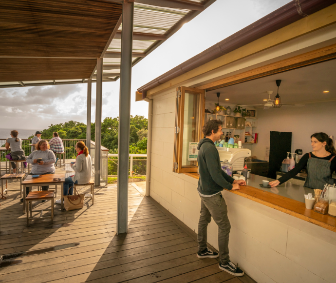 Coastal cafe with outdoor deck, serving customers overlooking ocean and trees.