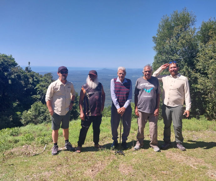 5 men on a high green clearing with a vista of forested plain visible low behind them and a blue sea on the horizon