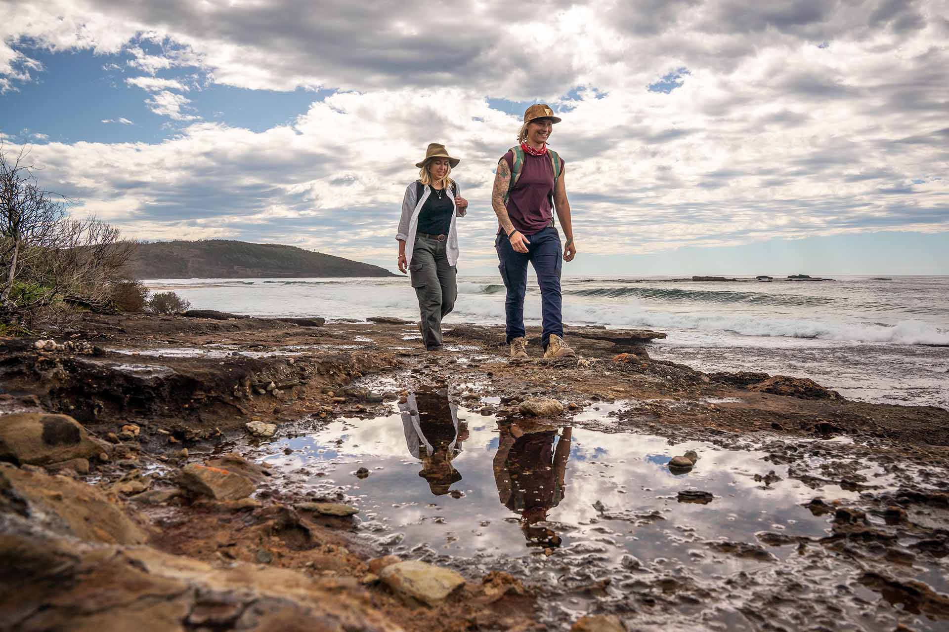 Two people in hiking gear walking past rock pools at the shoreline