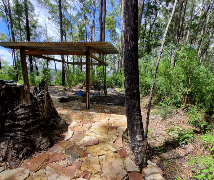 A simple, 4-post shelter standing on stone paving, at a high aspect among the trees with another hilltop visible in the distance