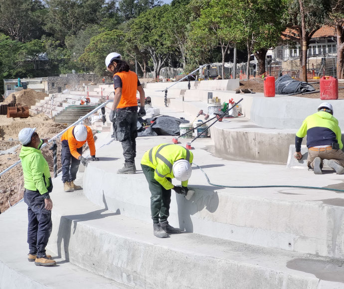 Five men in high visibility shirts and helmets work on some smooth new concrete bleachers with equipment and machinery behind them and on the sand; thick treeline in the background.