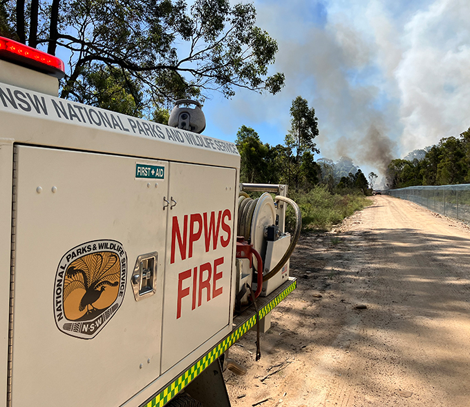 The image depicts a scene of a fire emergency in New South Wales, Australia. In the foreground, there’s a NSW National Parks and Wildlife Service (NPWS) fire truck parked on a dirt road. The side of the truck is labeled “NPWS FIRE” and also has a “FIRST-AID” logo. Equipment, including hoses for firefighting, is mounted on the side of the truck. In the background, smoke rises into the sky, indicating an ongoing wildfire. The natural environment features trees lining both sides of the dirt road, and a fence runs along one side.