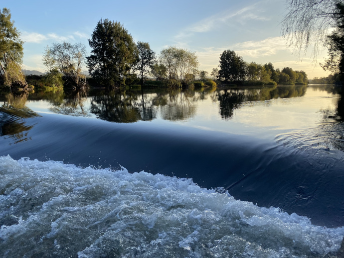 Foaming water of the the Hunter River in the foreground as it flows over Liddell Weir. Behind the weir the river surface is smooth, reflecting trees that line its edges in a golden light.