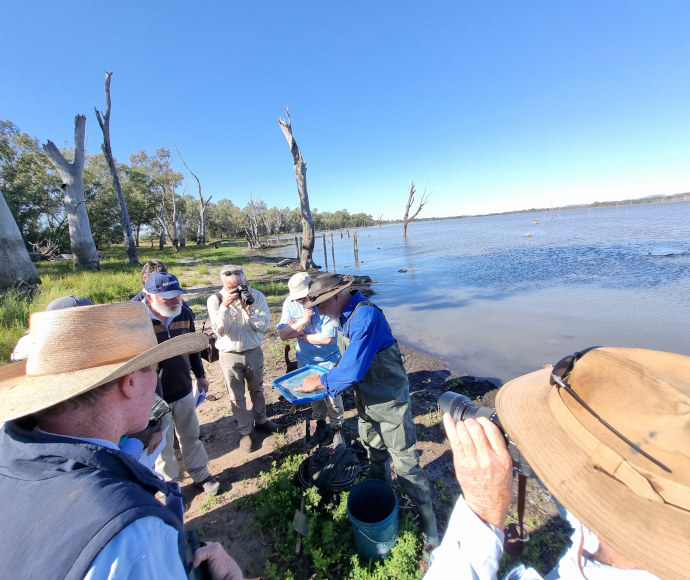 Gathered by the shore of a lake, a small group of people in sunsafe clothing - hats, long sleeves, long trousers - lean in to see a man presenting findings in a small container of water and organic matter. Leafless, branchless tree trunks stand like bones in the background.