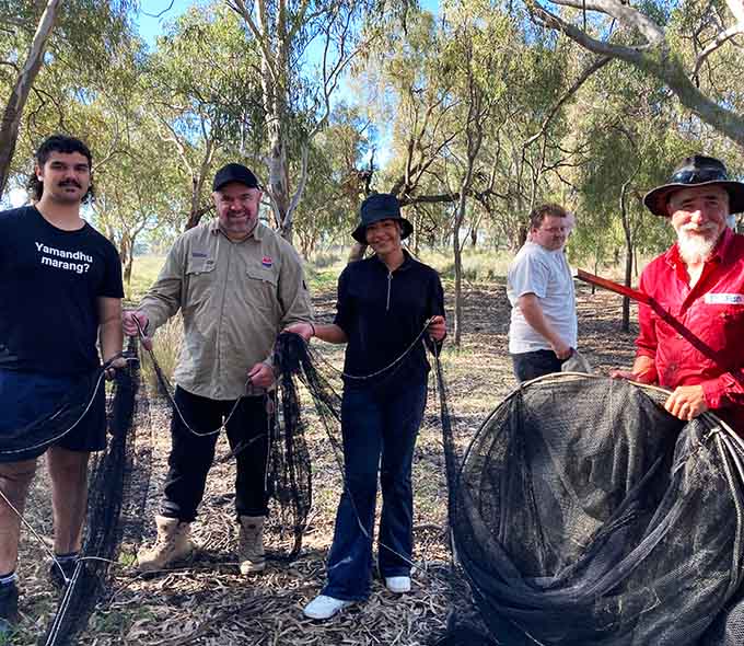 Five individuals stand outdoors holding large nets, with trees and vegetation in the background